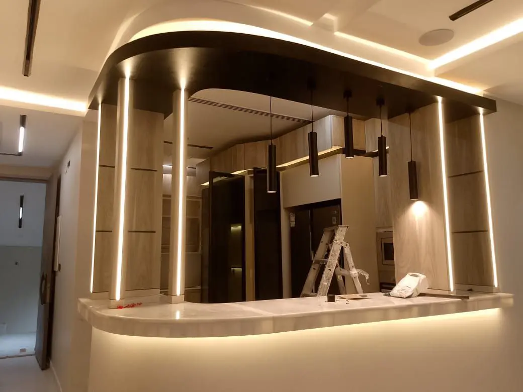 صورة تبرز تنفيذ إنارة بروفايل ليد (Profile LED) رأسية وأفقية مدمجة بديكور خشبي ورخامي، مع إضاءة ستريب لايت مخفية تضفي لمسة جمالية دافئة وتصميم مدروس للمساحات الحديثة.